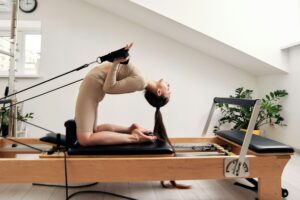 A young girl is doing Pilates on a reformer bed in a bright studio. A slender brunette in a beige